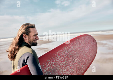 Man holding a full size surf board standing on beach looking out to sea Stock Photo