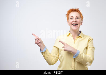 Surprised funny mature woman in yellow shirt showing empty copy space with her finger grey background. Just look on this proposal Stock Photo