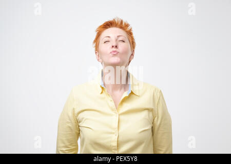 Portrait of cute lovely mature woman in casual yellow outfit with modern red colored hairdo sending blowing kiss with pout lips looking at camera isol Stock Photo