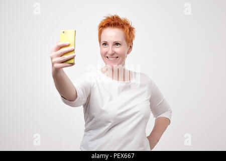 Pretty mature happy woman with red hair in white shirt making selfie on smartphone Stock Photo