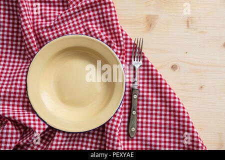 Rustic place setting. Empty enamel plate and fork, red checkered tablecloth on wooden table, top view, copy space Stock Photo
