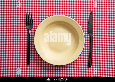 Rustic place setting. Empty enamel plate, fork and knife on red checkered tablecloth, top view Stock Photo