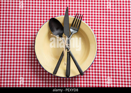 Rustic place setting. Empty enamel plate and cutlery on red checkered tablecloth, top view Stock Photo