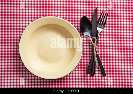 Rustic place setting. Empty enamel plate and cutlery on red checkered tablecloth, top view Stock Photo