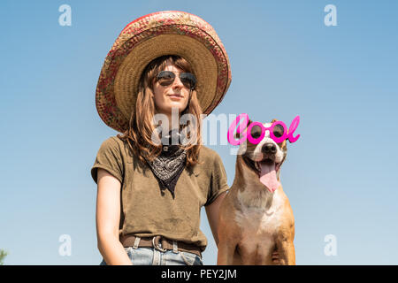 Beautiful girl in mexican hat dressed up as bandit of gangster with dog in cool sunglasses. Female person in sombrero hat and bandana posing with pupp Stock Photo