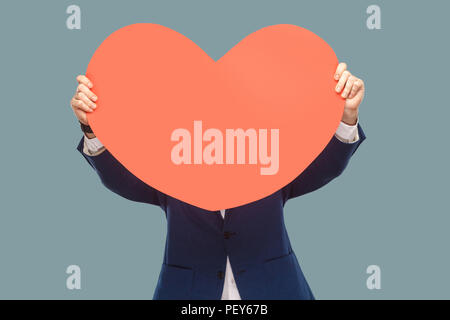 Handsome businessman in blue jacket standing and holding and covering red big heart shape in front of face for copy space. Indoor, studio shot isolate Stock Photo