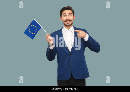 Handsome businessman in blue jacket and white shirt holding and pointing finger at European union flag and looking at camera with toothy smile. Indoor Stock Photo