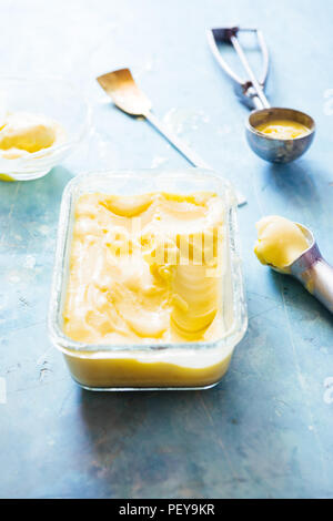 Mango ice cream on a glass container with icecream utensil, on a blue board, frontal view with natural back light Stock Photo