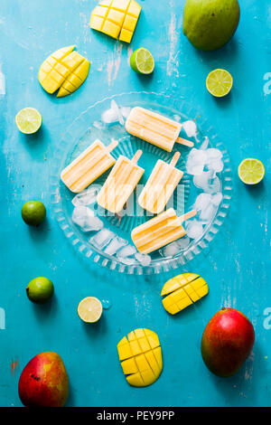 Mango popsicles on a glass plate with ice cubes, lime and mango fruit,top view, blue board. Stock Photo