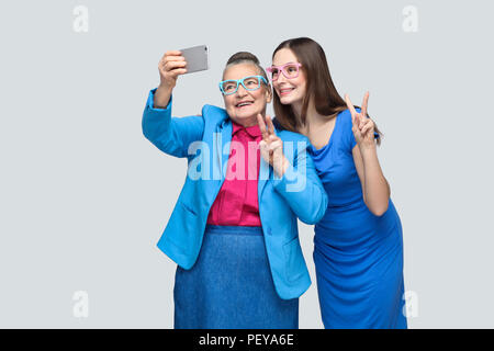 happy grandmother with granddaughter in blue dress or suit toothy smiling, standing, posing and making selfie and peace sign. Relations in the family. Stock Photo