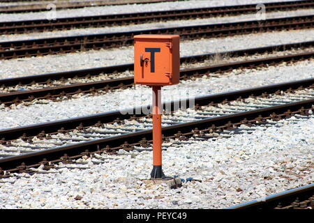 Railway trackside orange communication utility box with lock and key mounted on metal pole and positioned on gravel between railway tracks on warm sun Stock Photo