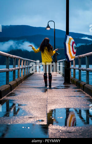 Young woman in a yellow raincoat standing under an umbrella on a rainy day by the ocean. Stock Photo