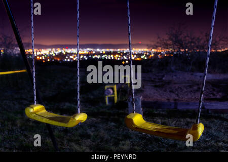 Empty swings on the night with city lights blurred on the background in a winter night Stock Photo