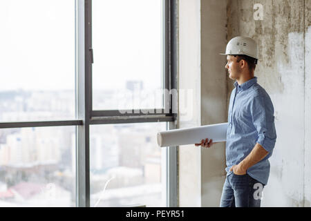 handsome guy in hardhat putting his hand on the pocket and looking at the panorama window. side view photo Stock Photo