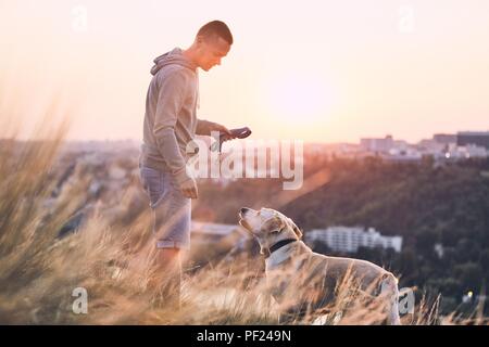 Morning walk with dog. Young man and his labrador retriever on the meadow against city at sunrise. Stock Photo