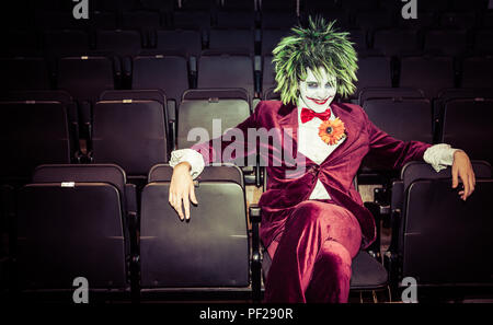 A male cosplayer dressed as The Joker from the Batman and DC Comics franchise sitting alone in an abandoned movie theatre and looking menacing Stock Photo