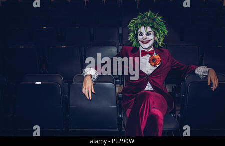 A male cosplayer dressed as The Joker from the Batman and DC Comics franchise sitting alone in an abandoned movie theatre and looking menacing Stock Photo
