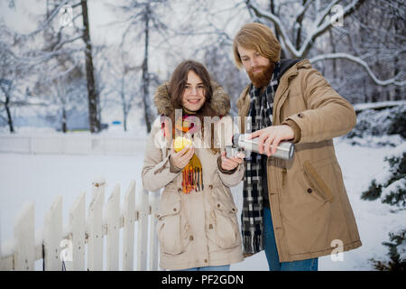 Young loving couple caucasian man with blond long hair and beard, beautiful woman have fun drinking a hot drink from thermos, eating green apple in sn Stock Photo