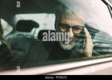Smiling senior businessman making phone call while sitting on back seat of a car. Senior male commuter travelling by a cab and talking on phone. Stock Photo