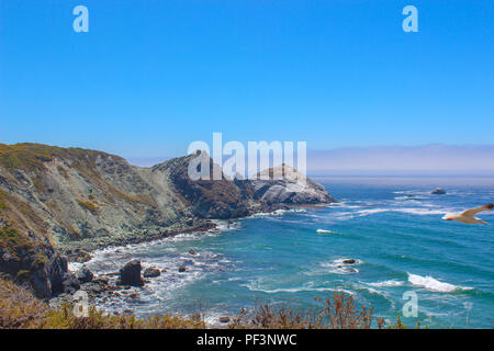 View looking West over a beautiful bay with an Azure Sea and clear Blue Sky, White tipped waves crash against the rugged rocks surrounding the bay. Stock Photo