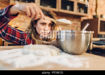 Young housewife in an apron puts flour in a bowl, kitchen interior on background. Female cook prepares fresh homemade cake. Domestic pie preparation Stock Photo