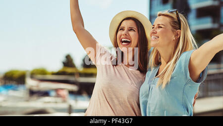 Two laughing young female friends standing arm in arm while having a good time together in the city in summer Stock Photo