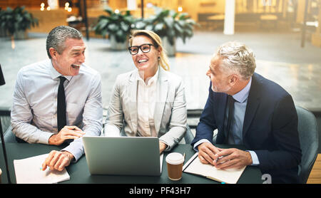 Three corporate colleagues laughing and talking together over a laptop at a table in the lobby of an office building Stock Photo