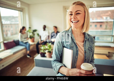 Smiling young businesswoman standing in an office carrying a laptop and holding a coffee with colleagues sitting in the background Stock Photo