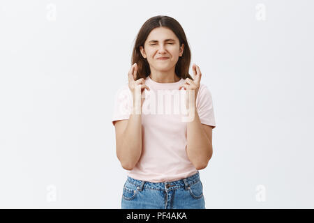 Good-looking european girl putting all effort in wishing for good luck while crossing fingers and closing eyes smiling broadly having faith that dream come true while standing over gray background Stock Photo