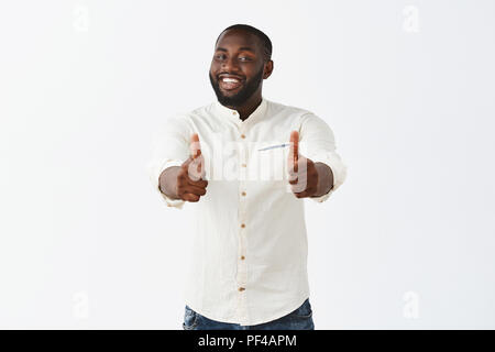 Studio shot of pleased good-looking crafty male african american chef in white shirt, smiling broadly from satisfaction, showing thumbs up at camera, liking new meal and suggesting everyone try it Stock Photo