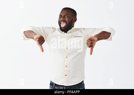 Studio shot of emotive joyful funny dark-skinned adult man with beard sticking out tongue and showing dislike, being unimpressed, making thumbs down with both hands and grimacing from disappointment Stock Photo