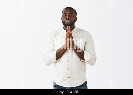 Indoor shot of serious-looking focused african american male with beard and short haircut holding hands in pray over chest in asian style, staring at camera, making namaste gesture over gray wall Stock Photo
