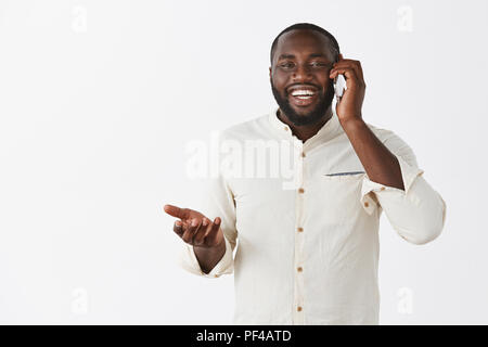 Charming carefree and happy man retelling amusing and funny story via smartphone, gesturing with hand while holding cellphone near ear and smiling broadly at camera over grey wall Stock Photo
