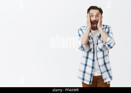 Man shouts seeing shocking situation, feeling nervous and scared. Intense displeased and anxious male with beard in checked shirt, yelling with hands in cheeks, being shocked and feeling desperate Stock Photo