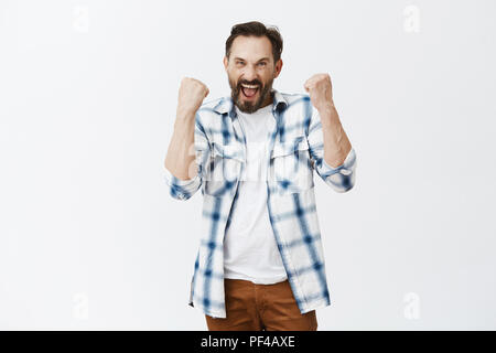 Excited dedicated football fan triumphing joyfully, raising clenched fists and yelling yes, enjoying great match and win of team, posing in casual outfit over gray background, celebrating success Stock Photo