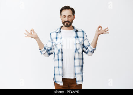 Stress goes away with meditation. Portrait of charming relaxed and calm european man with beard and moustache in checked shirt, raising hands aside in zen gesture, closing eyes and practicing yoga Stock Photo