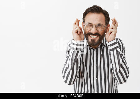 I have to win. Excited and nervous adult man with beard in formal clothes and glasses, raising fingers crossed, clenching teeth, making wish, feeling worried while waiting for good news Stock Photo