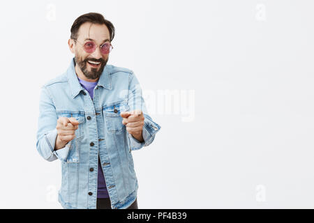 Your turn to buy me drink. Portrait of charming cheerful and carefree adult caucasian man with beard in sunglasses and trendy denim jacket, pointing at camera with index fingers and smiling broadly Stock Photo
