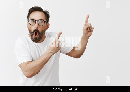 Studio shot of shocked and surprised impressed man with beard and moustache in trendy black glasses, pointing at upper right corner with both hands, dropping jaw and staring shook at camera Stock Photo