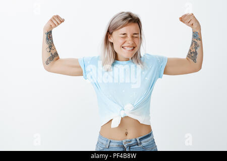 Girl power is strongest. Portrait of joyful good-looking active sportswomen with fair hair and tattooed arms, showing muscles or biceps, smiling broadly with closed eyes, being strong and confident Stock Photo