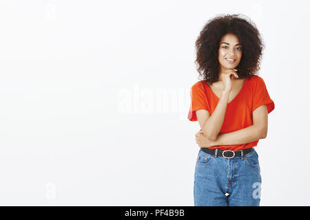 Studio shot of confident charming dark-skinned woman with afro hairstyle holding hand under chin and smiling broadly, crossing body with arm and gazing at camera, being interested in idea Stock Photo