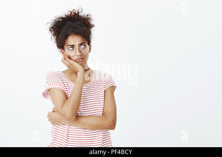 Girl feleing homesick and bored. Portrait of gloomy cute woman in glasses and striped t-shirt, leaning head on hand and looking up while frowning and sulking, being in bad mood over gray wall Stock Photo