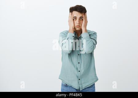 Portrait of depressed unhappy guy in trendy shirt, holding hands on head and staring with indifference at camera, suffering insomnia or stress, feeling tired and fed up over gray background Stock Photo