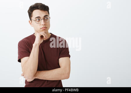 Portrait of smart creative male entrepreneur in t-shirt and round glasses, looking in right direction and thinking with half-crossed hands, holding palm on chin while making decision or solving issue Stock Photo