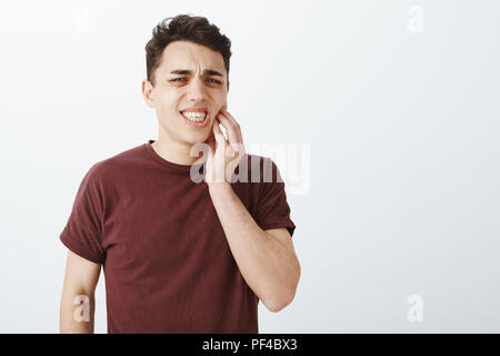 Portrait of displeased suffering young male model in red t-shirt, frowning and clenching fists, touching jaw while feeling toothache, having decay and dental problems, standing over gray background Stock Photo