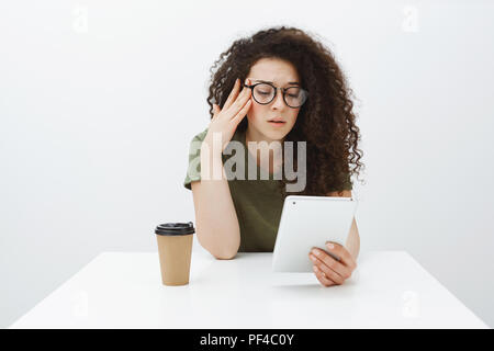 Tired fed up girlfriend with curly hair, sitting at table in cafe, drinking tea or coffee and reading article in digital tablet, holding hand on temple, being exhausted and unfocused over gray wall Stock Photo