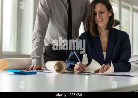 Woman working on a blueprint in an architects office as her partner or associate stands over her watching her in a low angle view across the table. Stock Photo