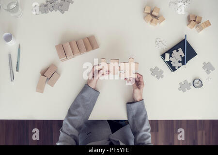 Top view of female hands assembling two wooden puzzle pieces with notebook, pen and other important metaphorical elements lying on white desk, toned r Stock Photo