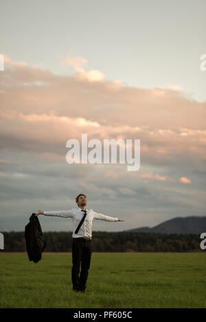 Success in business concept with a happy young businessman standing holding his jacket with outstretched arms in a rural field in evening light. Stock Photo
