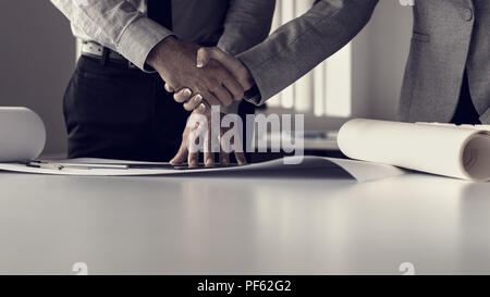 Retro toned image of businessman shaking hands with his female business partner analyzing blueprints in an office. Stock Photo